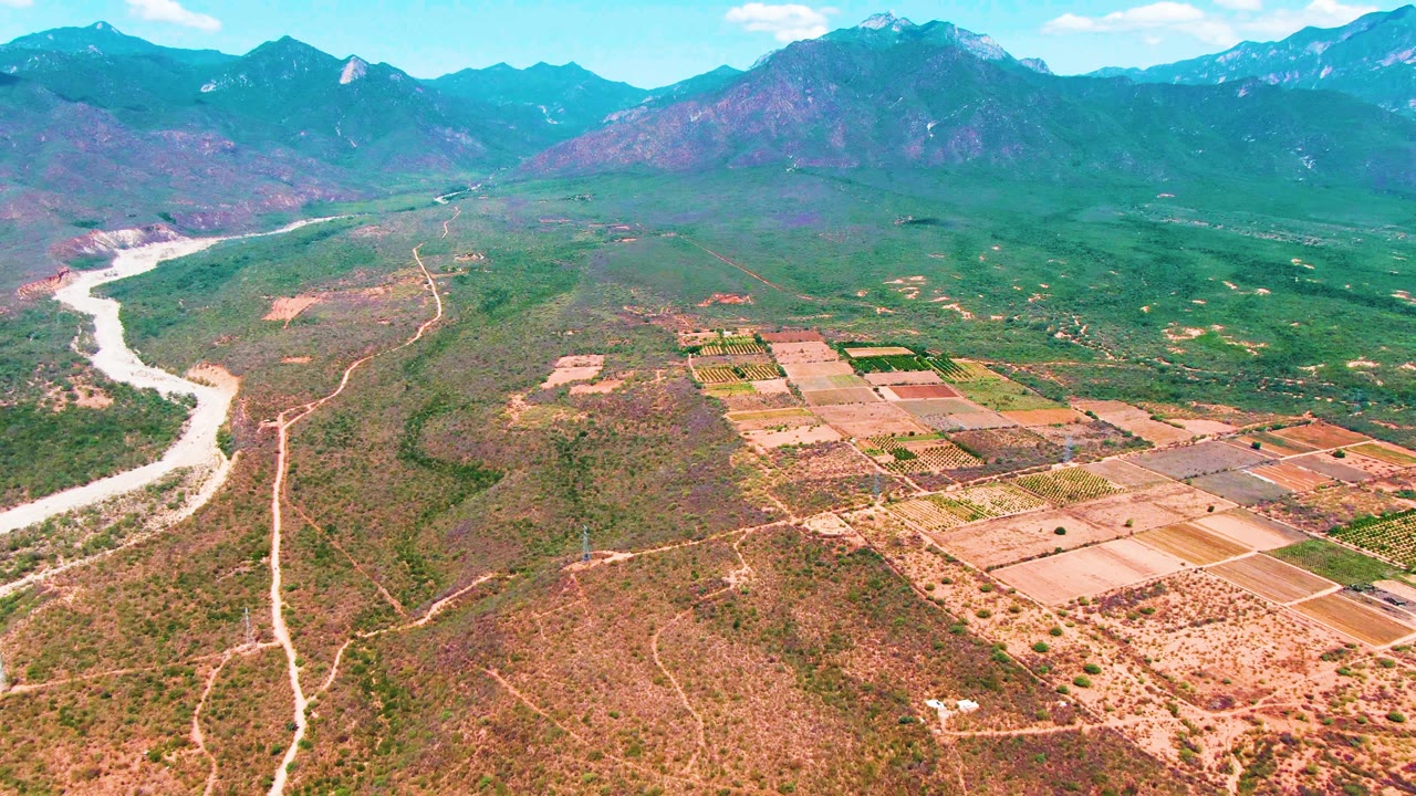 Aerial frame from Caduano, Baja California Sur, with the river curving along the left edge, farm parcels spreading across the right midground, dry valley floor in front, mountains behind, and no visible people.