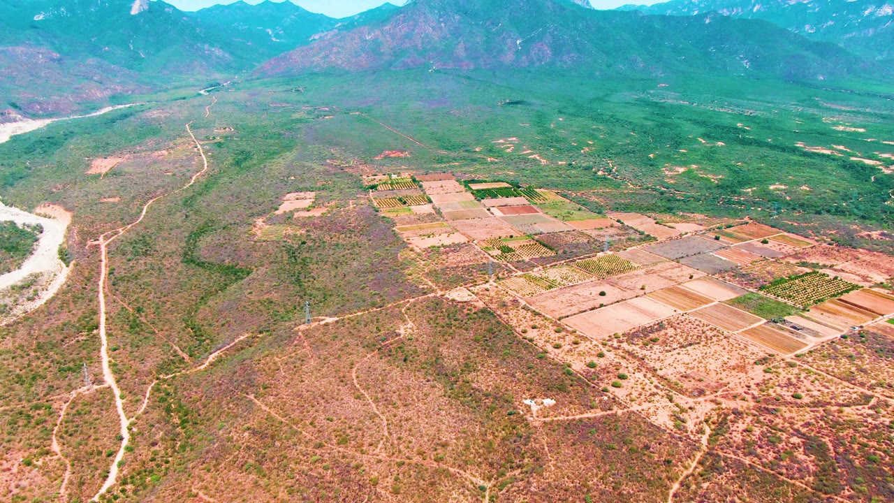 High aerial view over Caduano farmland with tan and green plots centered in the frame, branching dirt tracks across scrubland, a river channel along the left edge, and no visible people.