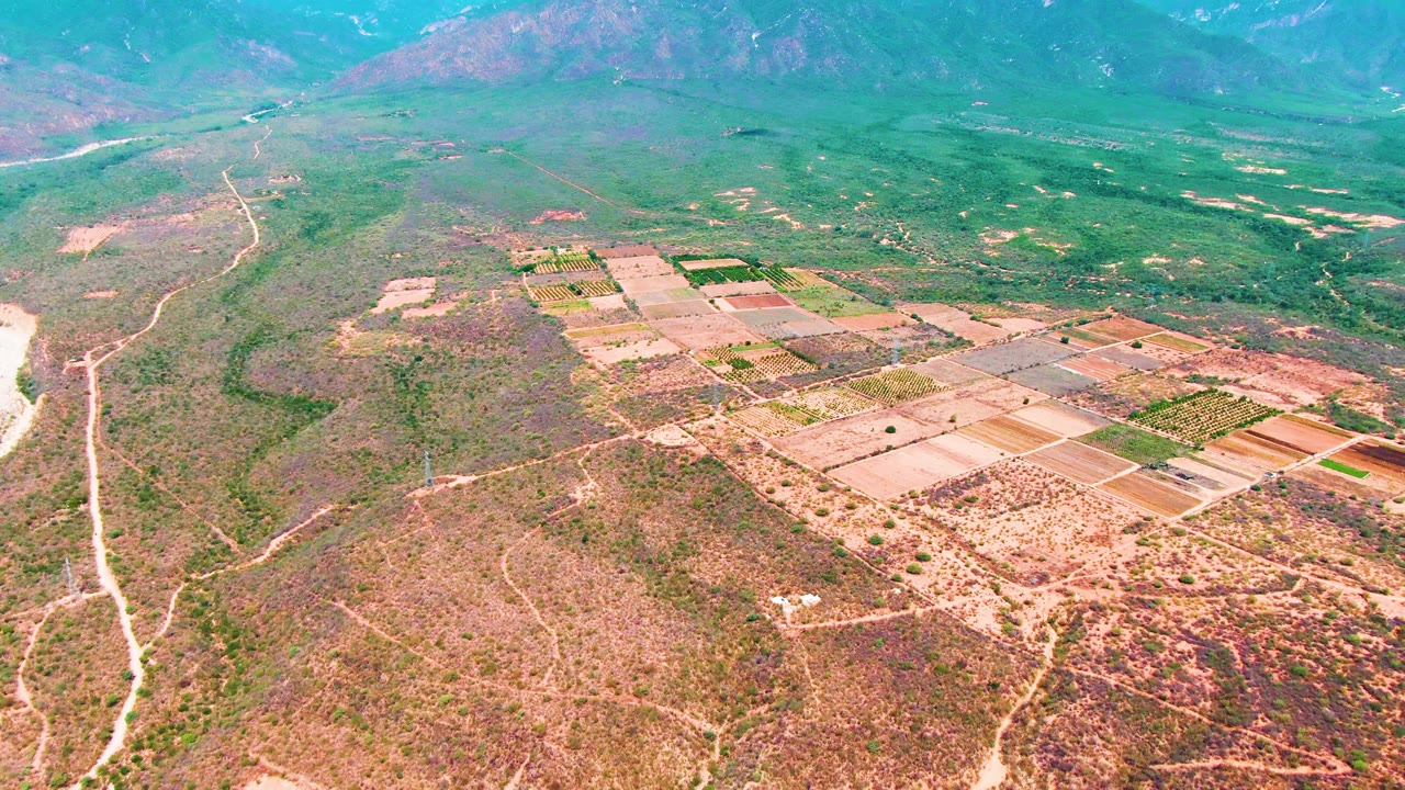 Aerial landscape over Caduano showing the farm grid lower right, dry brush and footpaths in the foreground, hazy green valley beyond, mountains on the horizon, and no visible people.
