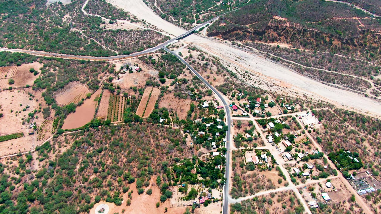 Drone view over Caduano with a paved road crossing a pale dry wash, rectangular fields on the left, scattered homes at lower right, and no visible people.