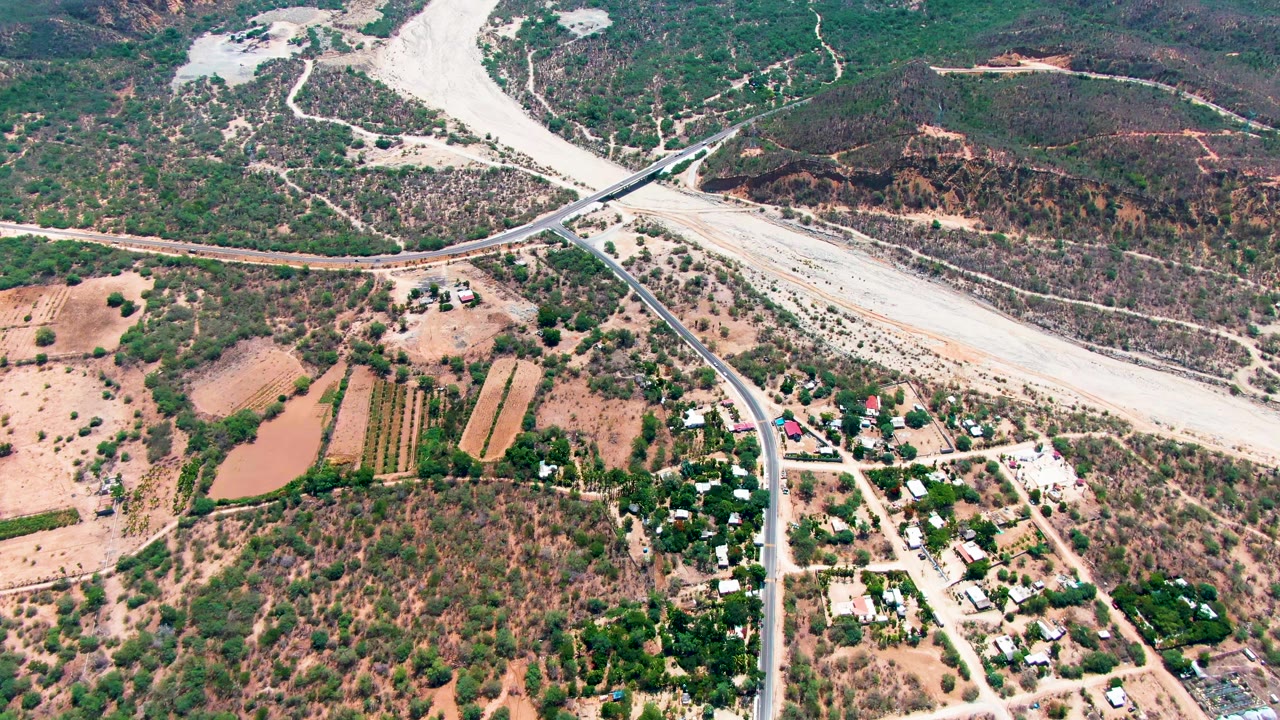 Aerial view over Caduano, Baja California Sur, with a pale riverbed crossing the upper frame, a bridge at center, fields and buildings below, and no visible people.