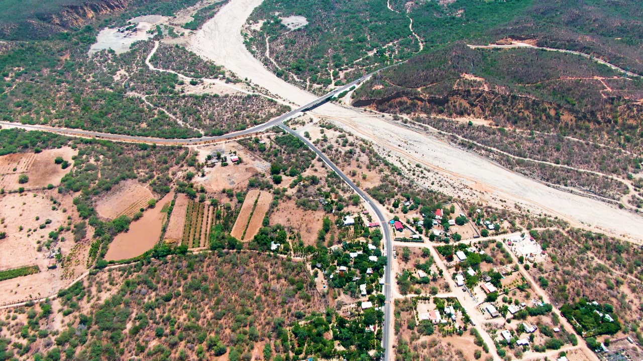 Second aerial frame over Caduano showing the bridge aligned near the center, pale river channel above it, agricultural plots and ponds below, and no visible people.