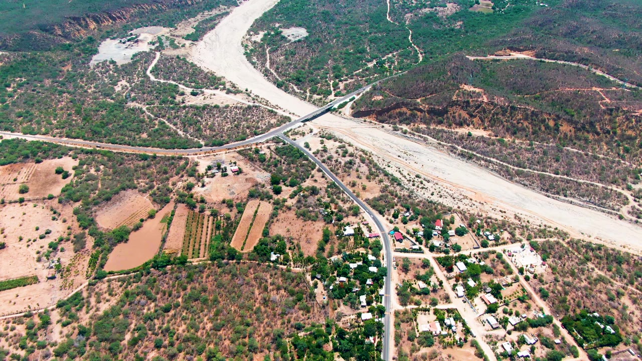 Aerial frame from Caduano, Baja California Sur, showing the pale riverbed spreading across the upper landscape, a bridge and road network, farmland, ponds, and no visible people.