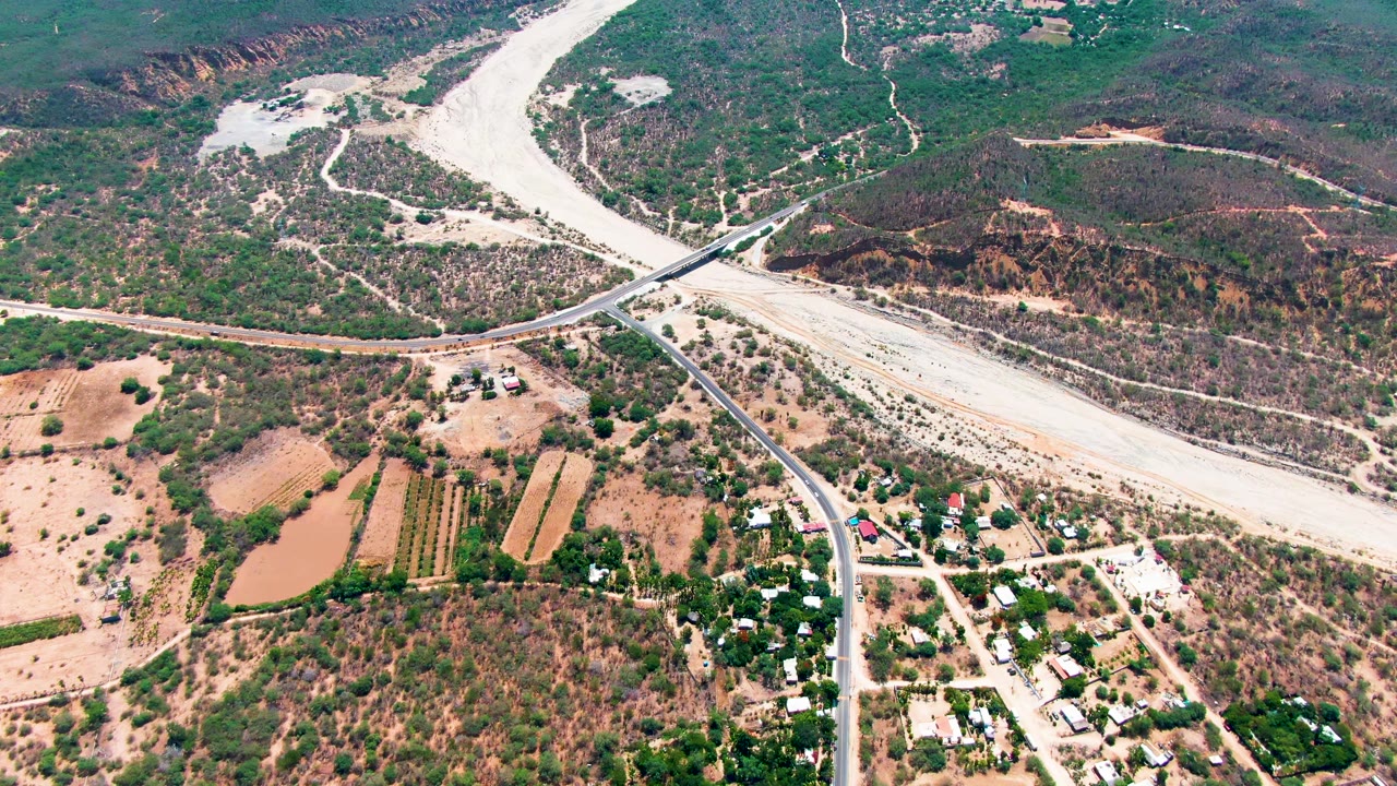 Aerial landscape with a bridge over a pale riverbed, rural roads, ponds, small fields, buildings, and brush.