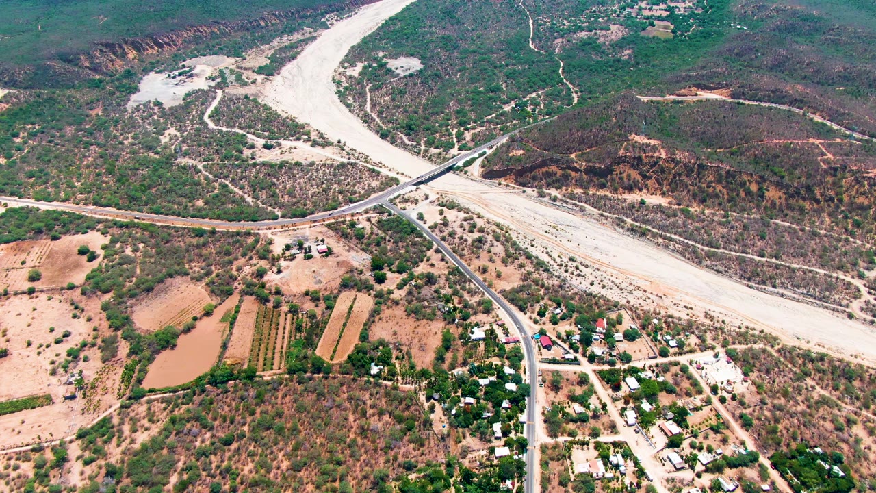 Aerial frame over Caduano, Baja California Sur, showing a pale dry riverbed crossed by a bridge, brown farmland, green shrubs, and small buildings with no visible people.