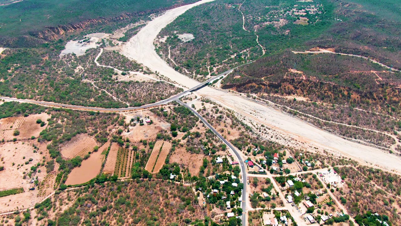 High aerial view over Caduano, Baja California Sur, showing the road bridge cutting across the dry river wash, rectangular farm plots, scrub-covered hills, and no visible people.