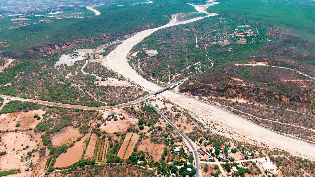 High aerial view over Caduano, Baja California Sur, showing a pale river channel, a road bridge, tan farmland, scattered buildings, green hills, and no visible people.