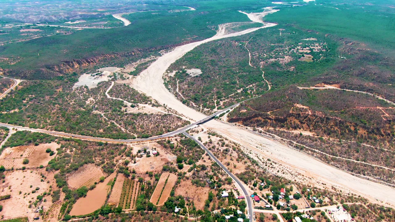 Aerial frame in Caduano showing branching pale floodwater channels curving through green scrub hills, with a road bridge and dry farm plots below, and no visible people.