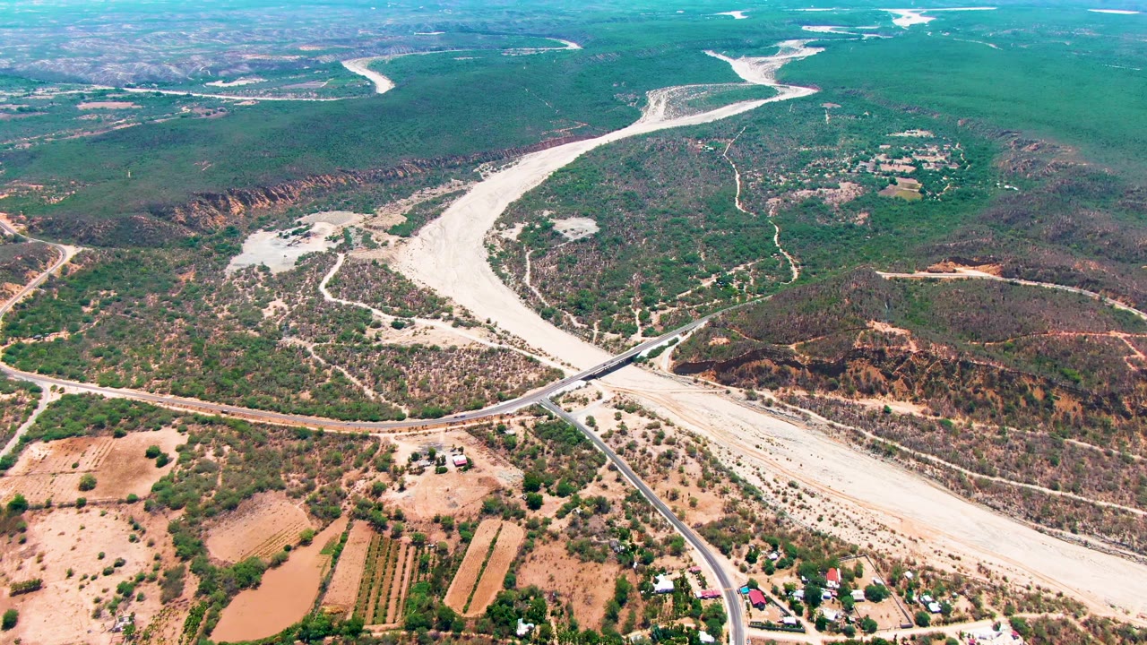Wide drone view over Caduano, Baja California Sur, with the bridge angled across the pale riverbed, tan fields at the bottom of frame, green hills beyond, and no visible people.