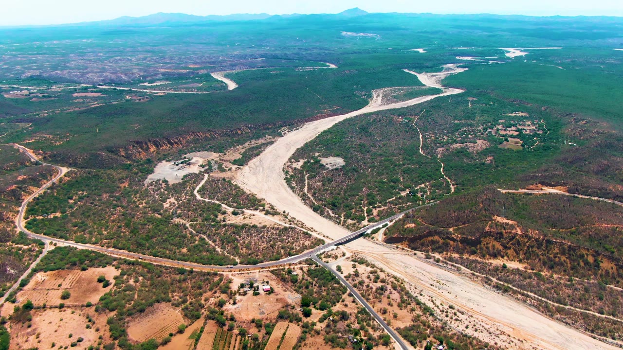 Aerial view of a pale riverbed winding through arid green-brown terrain with a bridge and distant mountains.