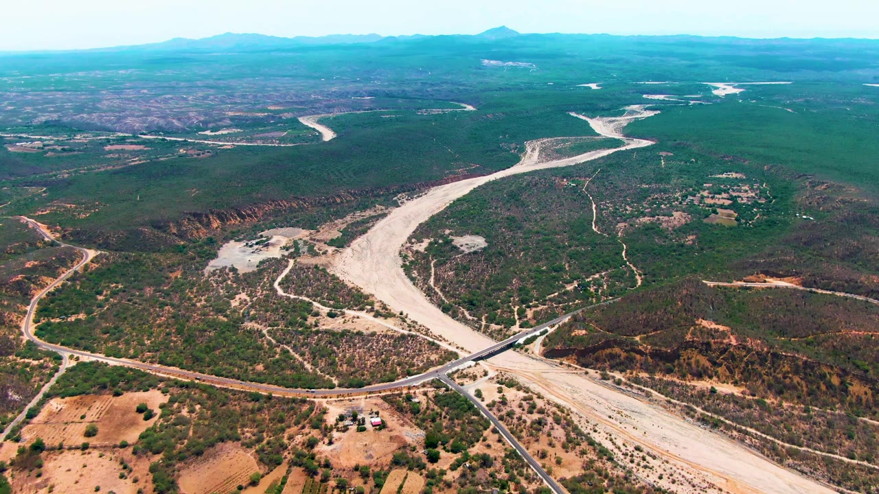 Wide drone view in Caduano, Baja California Sur, with the dry river channel centered, a bridge crossing near the bottom edge, patchy vegetation, distant mountains, and no visible people.
