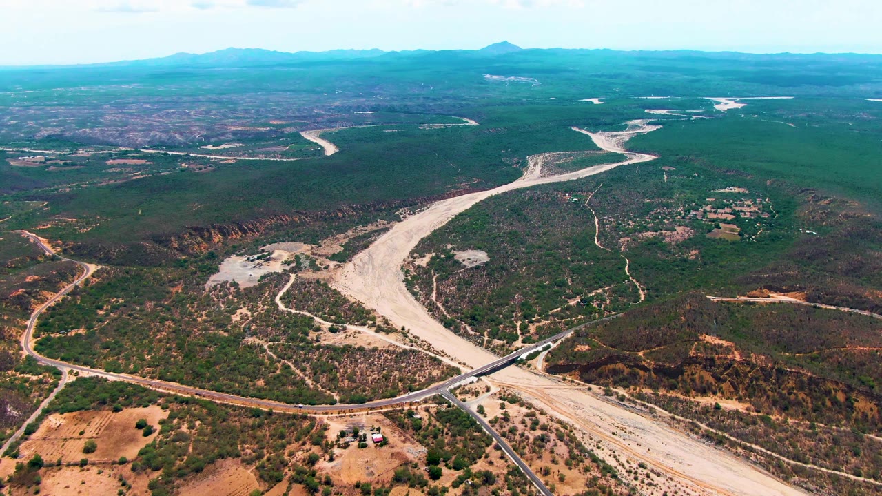 Aerial frame over Caduano, Baja California Sur, showing a branching pale riverbed through scrubland, rural roads in the lower terrain, a hazy mountain skyline, and no visible people.