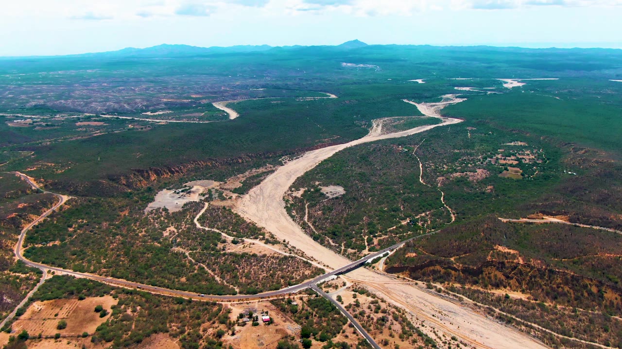 High aerial view in Caduano, Baja California Sur, with the pale riverbed curving past dark green hills, small tan fields, dirt tracks, a bridge at the bottom, and no visible people.