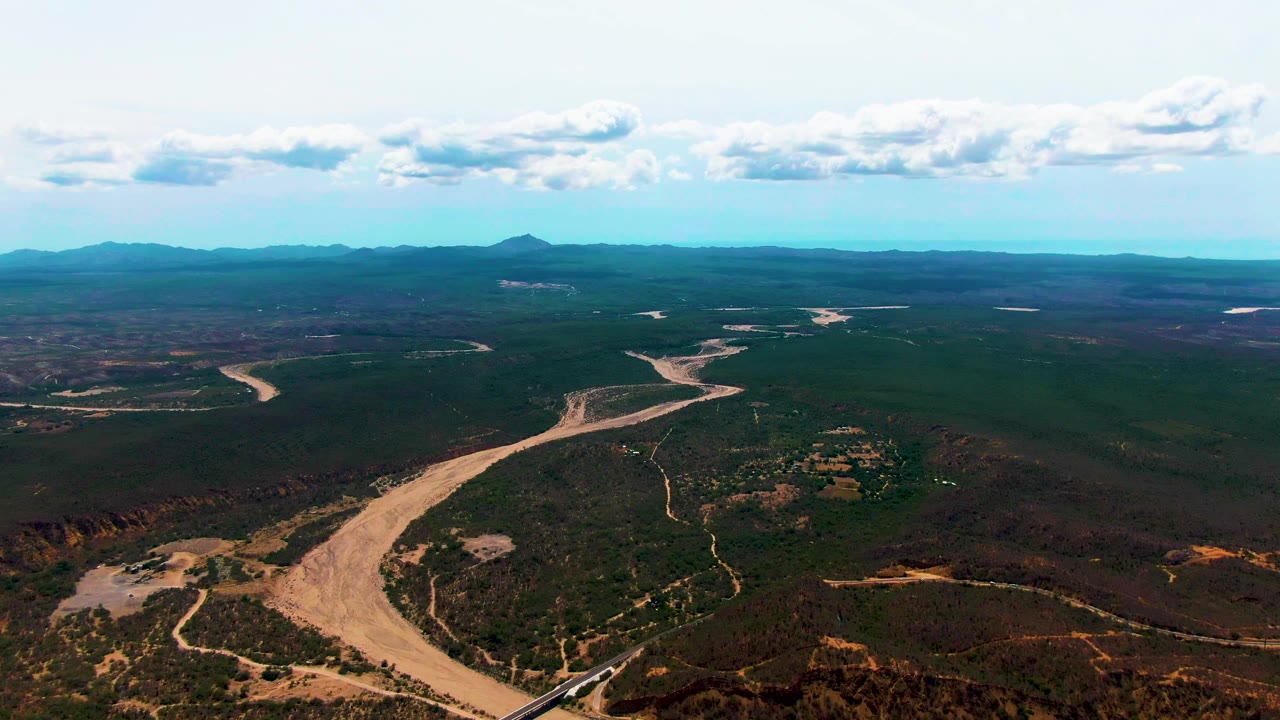 At 0:01, the Caduano aerial view holds on the broad river bends and rail corridor, with scrubland filling the midground, mountains along the horizon, bright clouds overhead, and no visible people.
