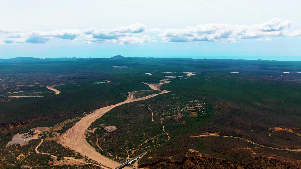 At 0:02, the pale river channel curves through Caduano terrain with branching dirt paths, a rail line at the lower frame, distant ridges, small cloud clusters, and no visible people.