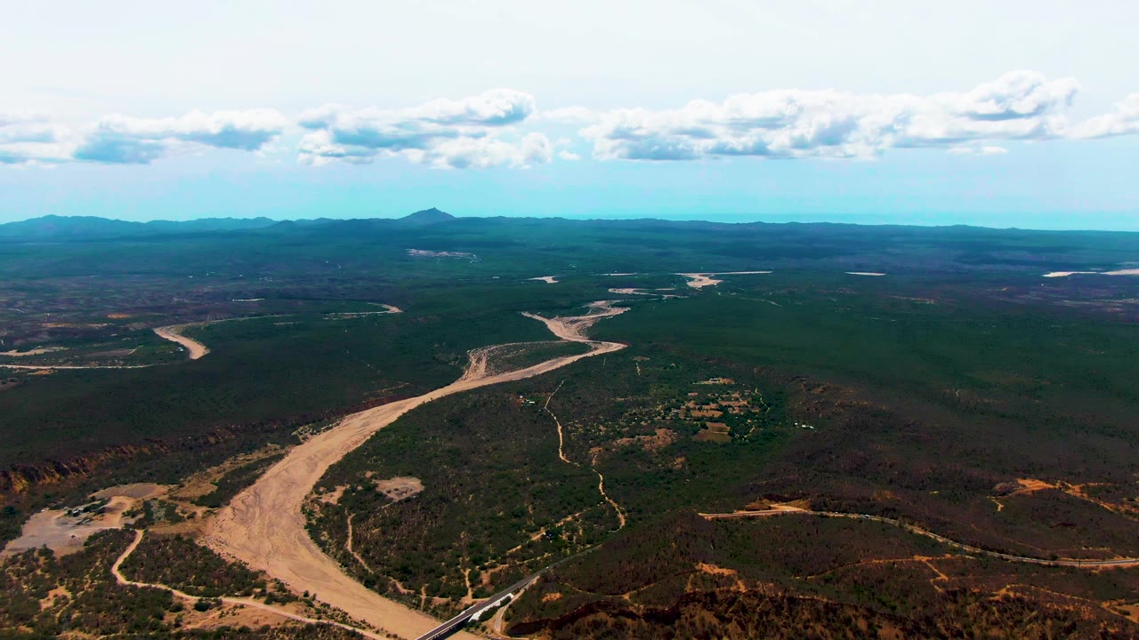 At 0:03, the wide aerial composition shows the winding river widening toward the lower left, the rail line near the bottom edge, open Caduano terrain, distant mountains, blue sky, and no visible people.