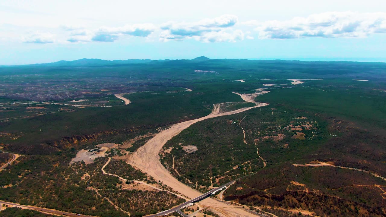 Aerial view of a winding riverbed, bridge, train, scrubland, mountains, and clouds.