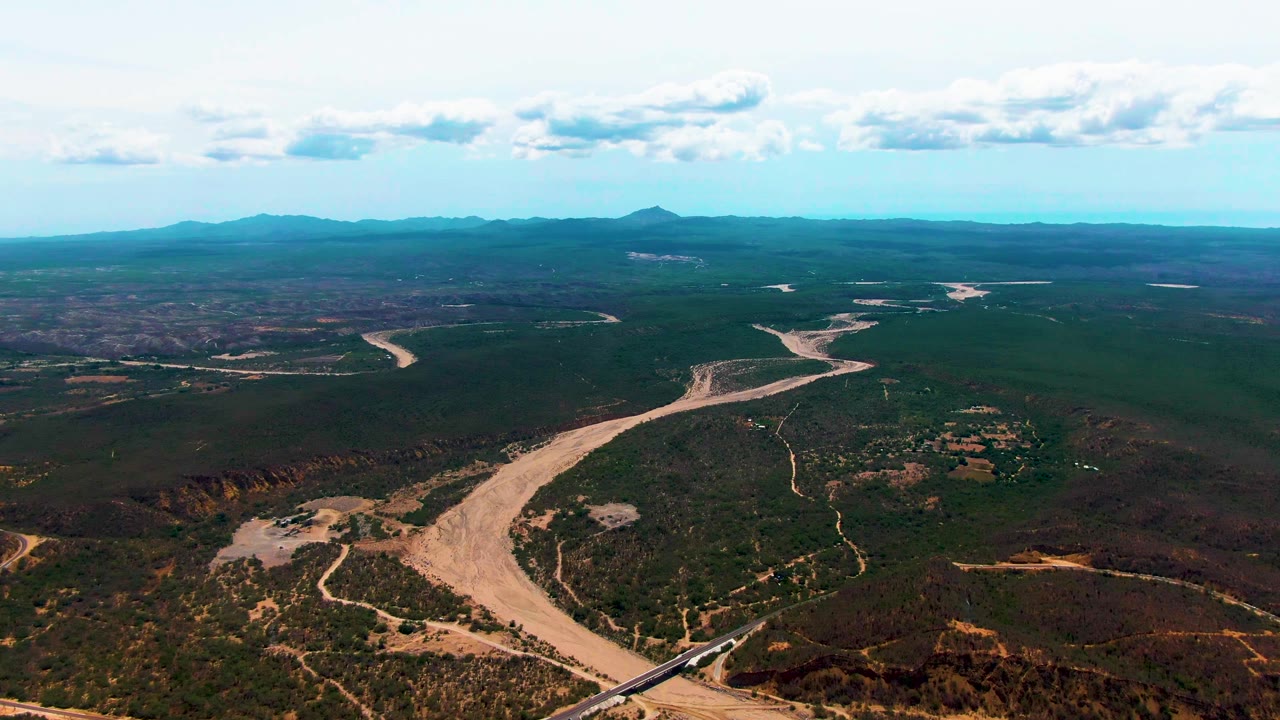 One second into the clip, the aerial view holds the Caduano river channel and bridge with a small train at the lower frame, scrub-covered terrain, distant hills, clouds, and no visible people.