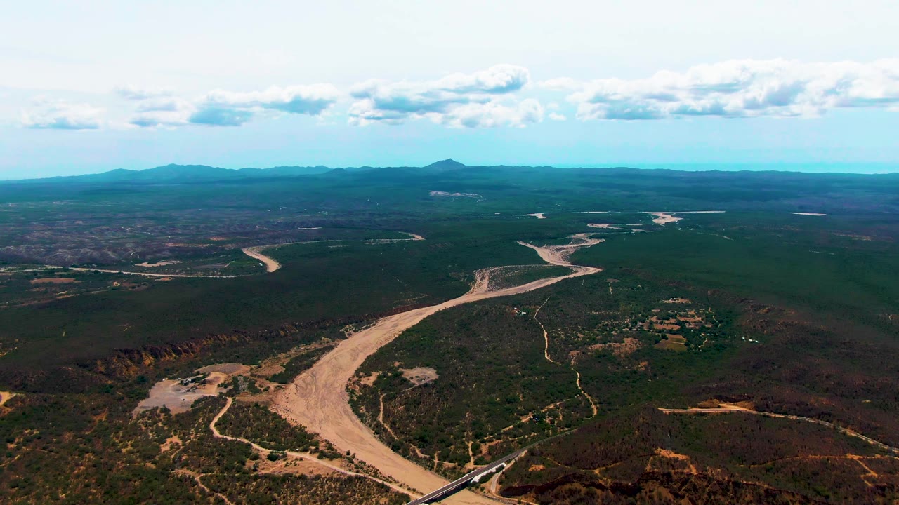 At 0:02, the Caduano aerial frame shows the pale riverbed sweeping through the center, the bridge and train along the lower edge, patchy vegetation, a broad horizon, and no visible people.