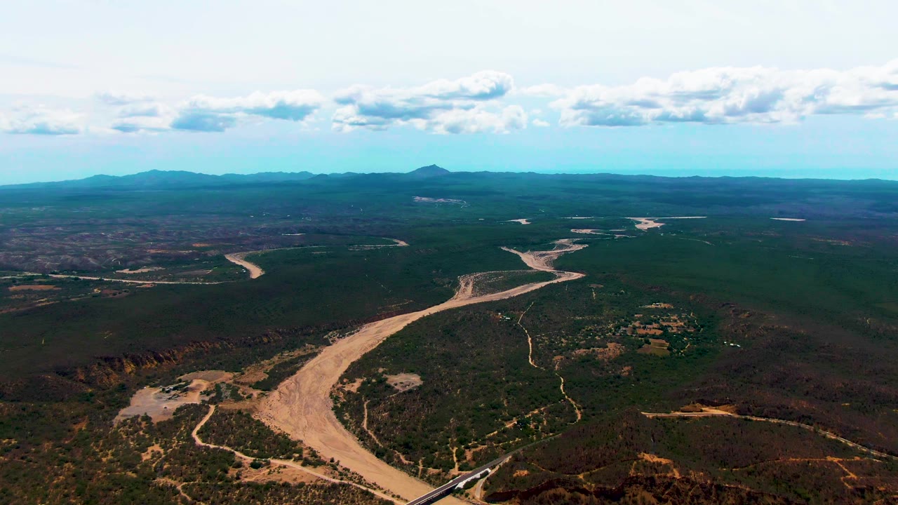 At 0:03, the Caduano landscape remains wide, with the bridge and train near the lower edge, branching dirt tracks, a winding riverbed, dark green scrubland, distant ridges, and no visible people.