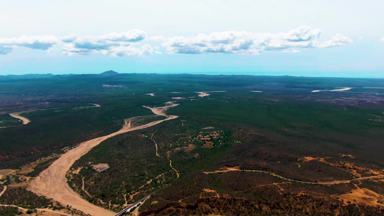 Wide aerial frame over Caduano, Baja California Sur, with a tan muddy river curving through dense green forest, dry brown tracks in the foreground, distant mountains, scattered clouds, and no visible people.