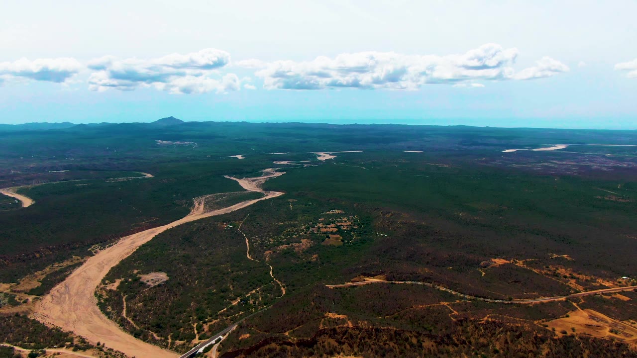Aerial frame in Caduano, Baja California Sur, showing the muddy river channel slightly farther along the valley, with dark green forest on both sides, dry foreground slopes, a low horizon, and no visible people.