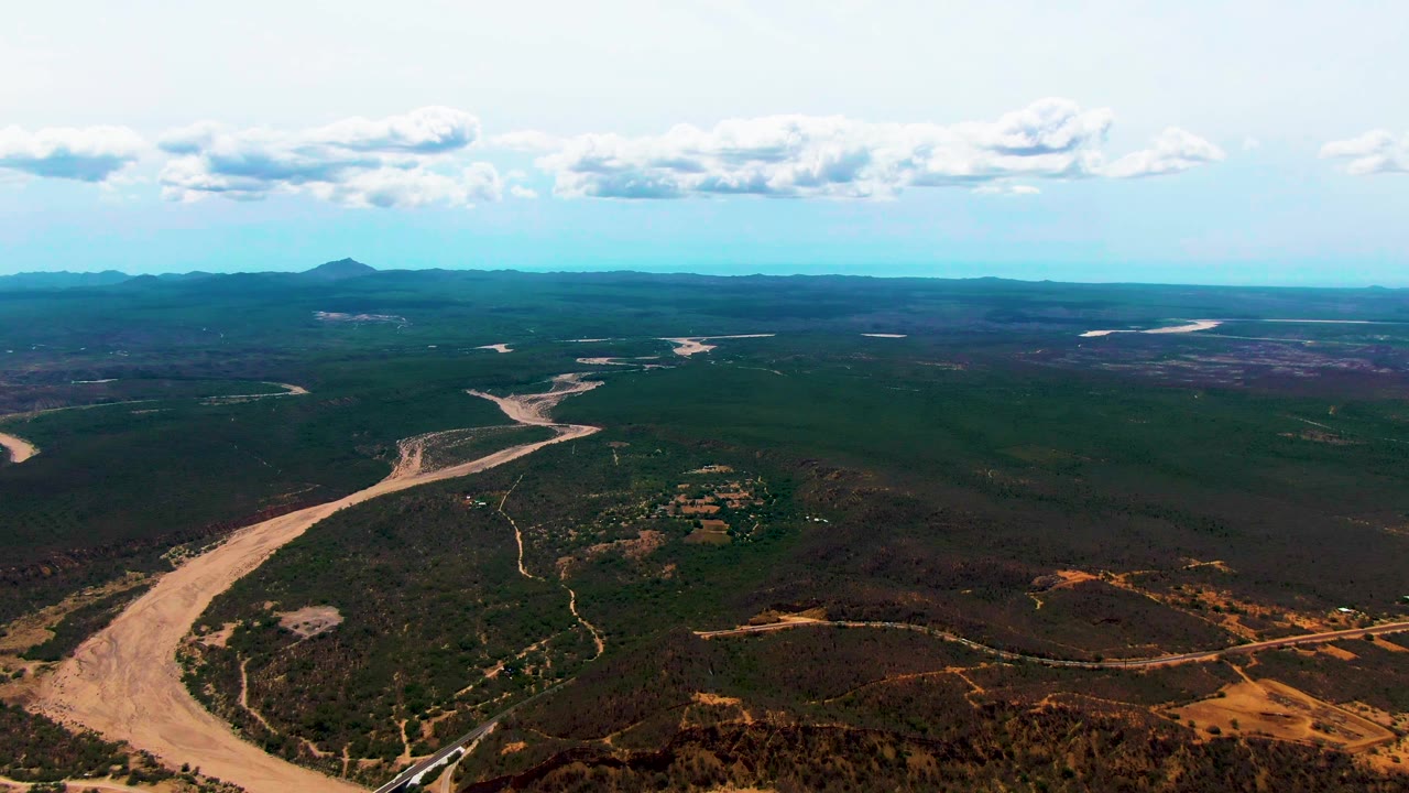 Wide Caduano aerial view with the tan river bending across the forested valley, small dry clearings and tracks below, hazy mountains along the horizon, scattered white clouds, and no visible people.