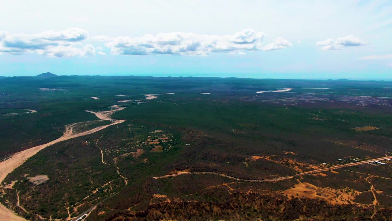 Aerial frame over Caduano showing the river channel nearer the center, a straight road across the lower right, scrub-covered ground, distant mountains, clouds, and no visible people.