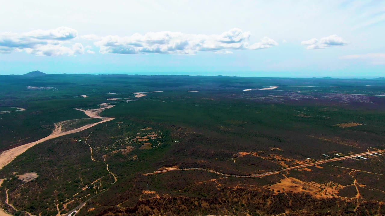 High aerial view in Caduano with a pale riverbed bending through dark green vegetation, brown patches in the foreground, a blue horizon, white clouds, and no visible people.