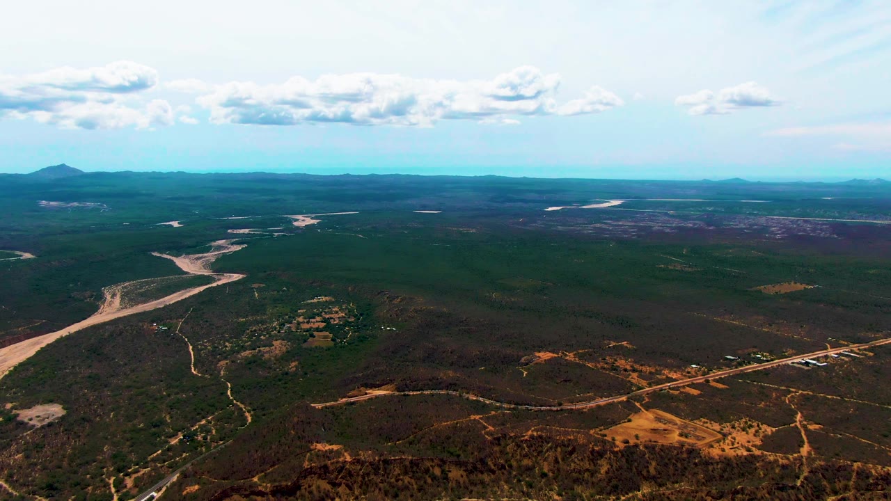 Wide drone view over Caduano with the river channel shifted left of center, a road running across the lower right, forested plain, distant ridges, scattered clouds, and no visible people.