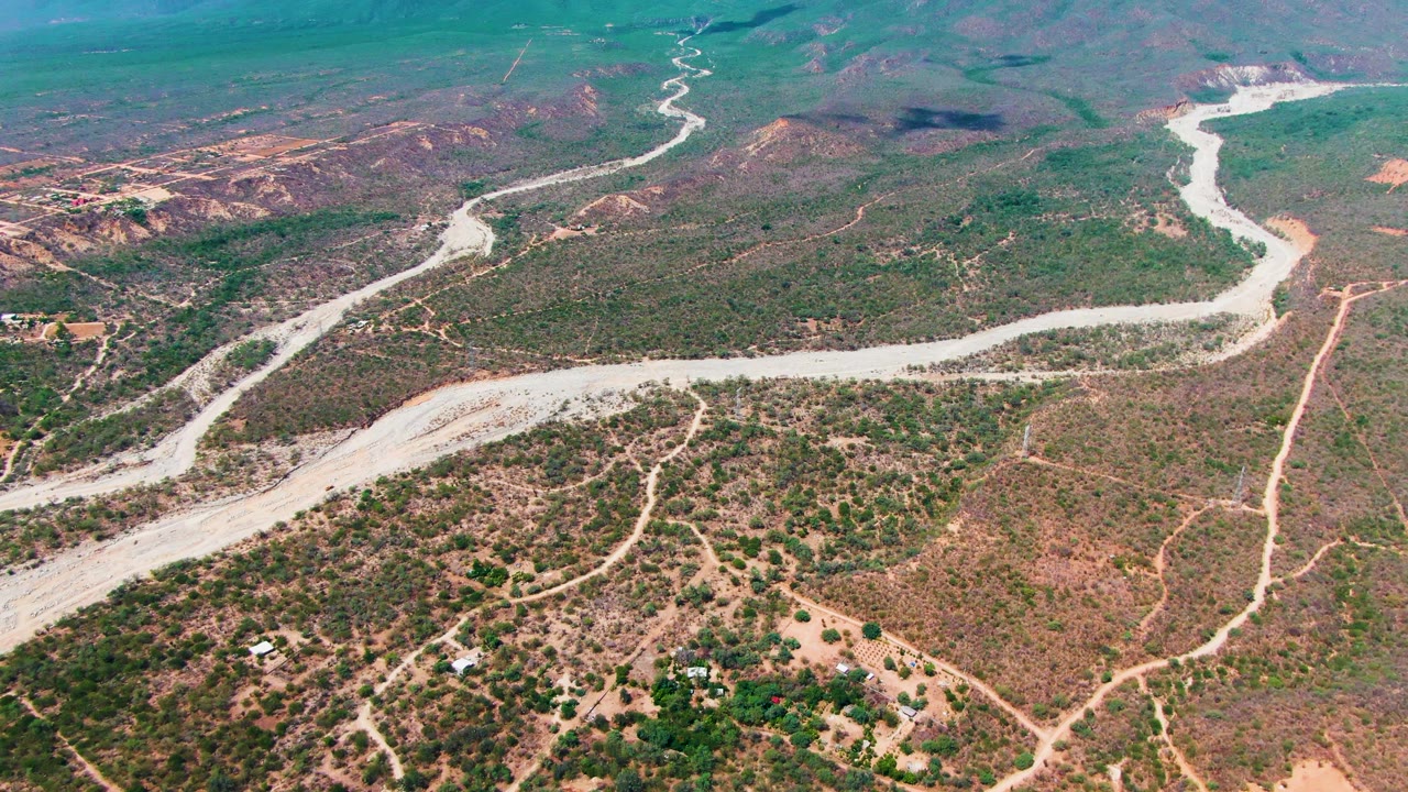 Aerial frame in Caduano, Baja California Sur with the pale winding corridor cutting across scrubland, dirt tracks branching through reddish hills, and no visible people.