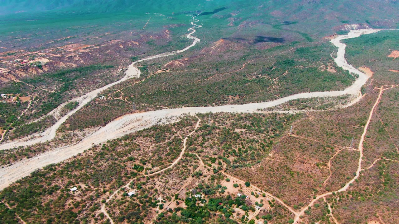 Wide aerial over Caduano, Baja California Sur showing the pale route bending through green-brown scrub, with branching dirt paths, low mountain ridges, and no visible people.
