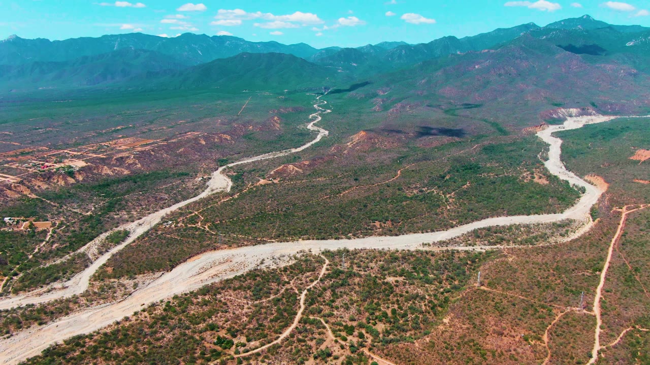Aerial frame over Caduano, Baja California Sur showing a pale winding road crossing scrubby brown and green terrain toward a mountain range, with no visible people.