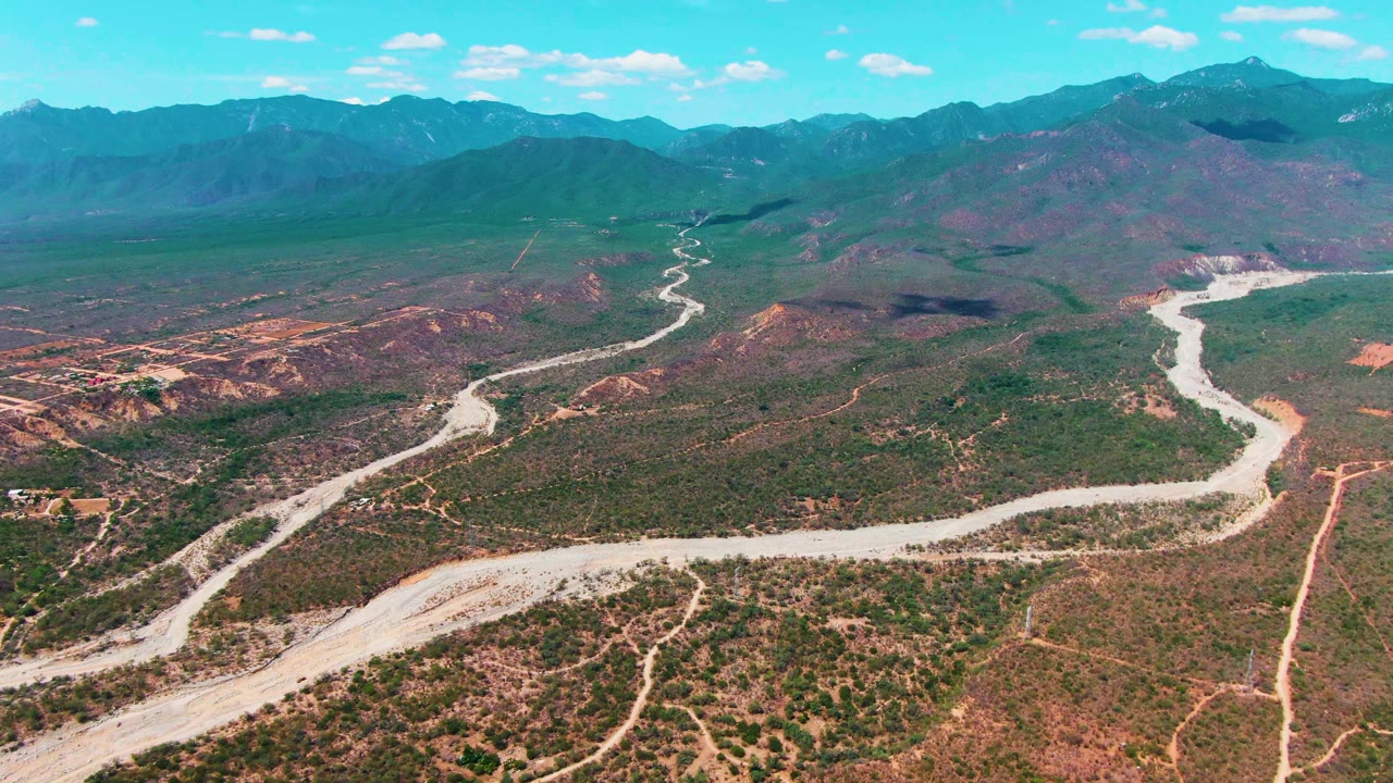 High aerial view in Caduano, Baja California Sur with the winding road centered through dry scrubland, reddish hills to the side, distant mountains, and no visible people.