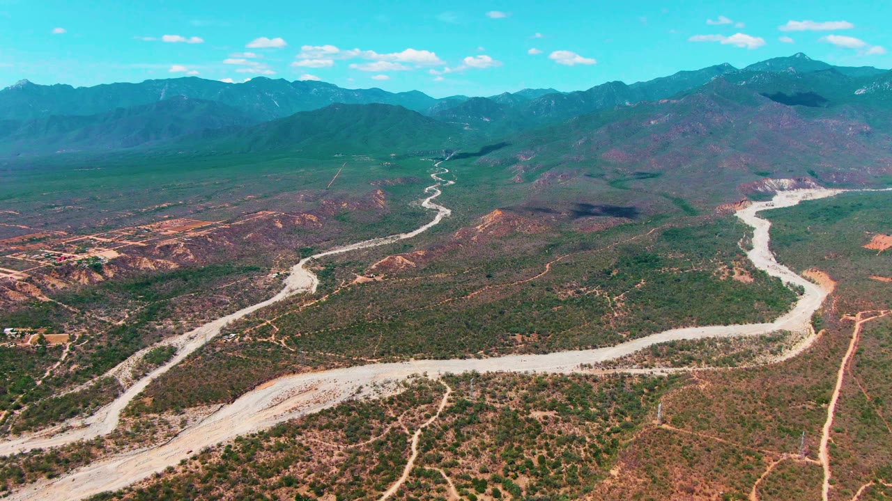 Wide drone frame from Caduano, Baja California Sur showing a pale road or dry wash snaking across the valley floor beneath layered mountain ridges, with no visible people.