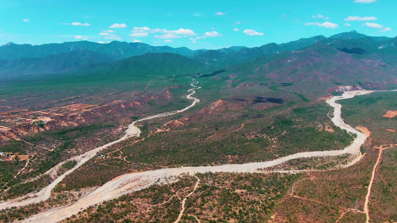 Aerial landscape over Caduano, Baja California Sur with a bright winding road cutting across sparse vegetation, darker green foothills, distant mountains, and no visible people.