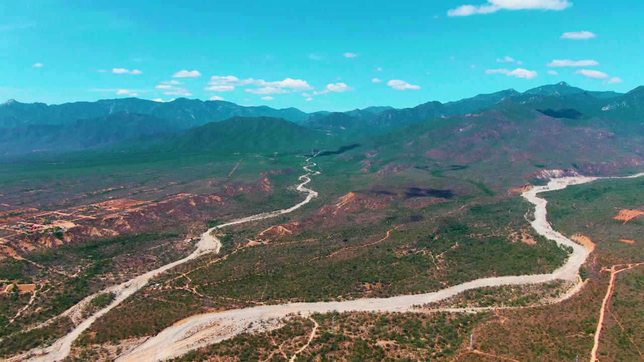 Wide aerial view of a winding pale road through scrubby mountain terrain.