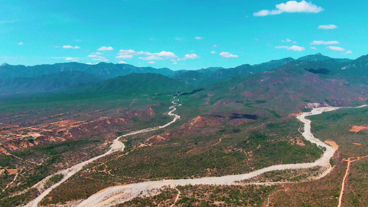 Aerial view in Caduano, Baja California Sur, with the winding road more centered through the valley floor, red dirt patches, mountain ridges, blue sky, and no visible people.