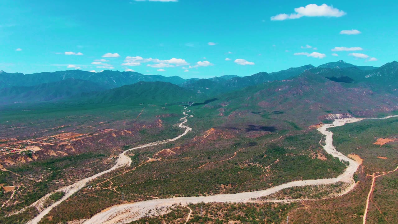 High aerial frame over Caduano, Baja California Sur, showing rugged low hills, a pale road bending from foreground to midground, distant mountains, scattered clouds, and no visible people.