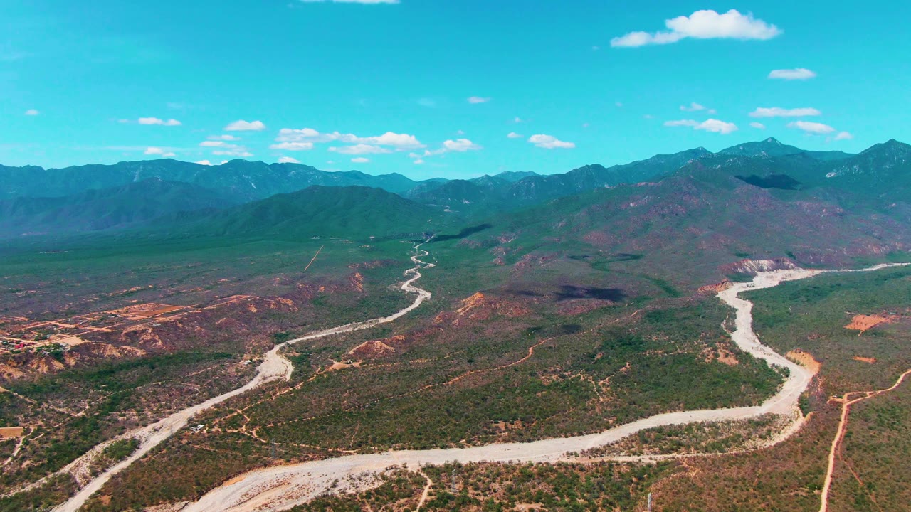 Elevated aerial frame in Caduano, Baja California Sur, with the road running diagonally through scrubland, red-brown ground exposed at left, mountain ridges behind, and no visible people.