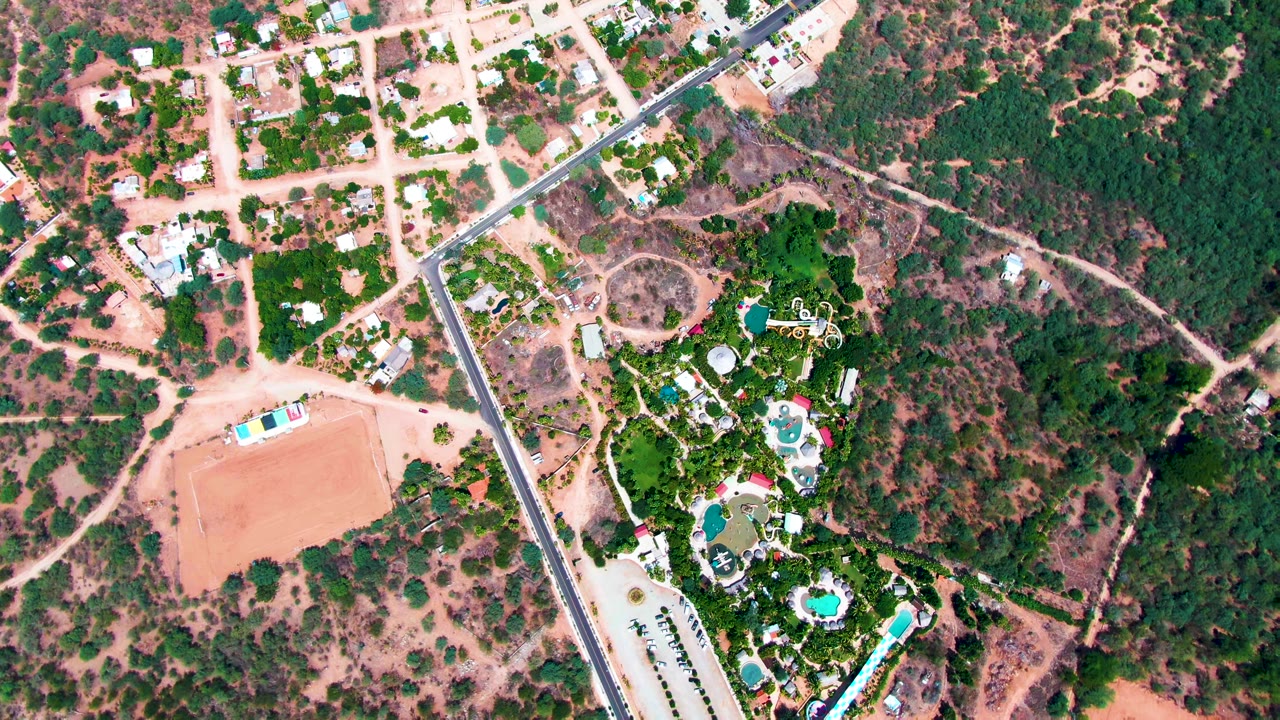 Overhead aerial frame in Caduano, Baja California Sur, showing a paved road, pale dirt lots, scattered homes, dense trees, and a landscaped estate with blue pools, with no visible people.