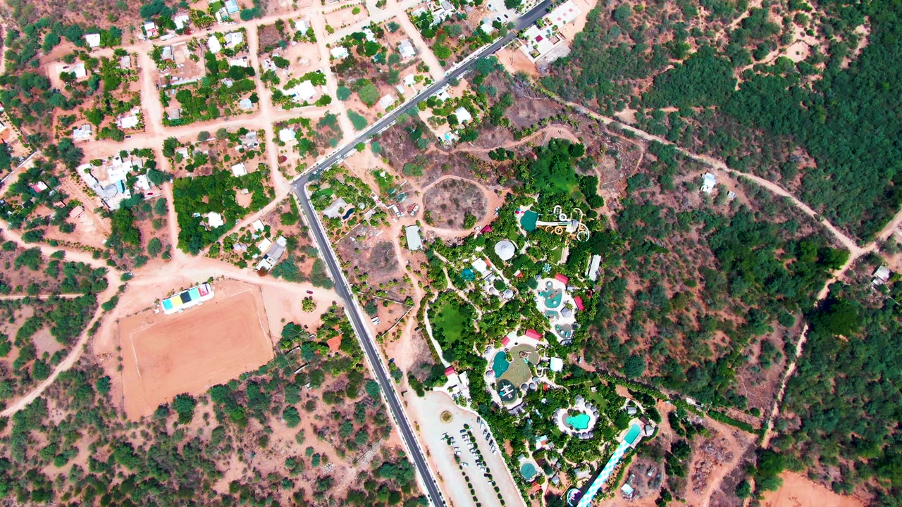 Top-down view in Caduano, Baja California Sur, showing clustered houses near pale roads, a band of dense green trees, reddish open ground, and multiple blue pools, with no visible people.