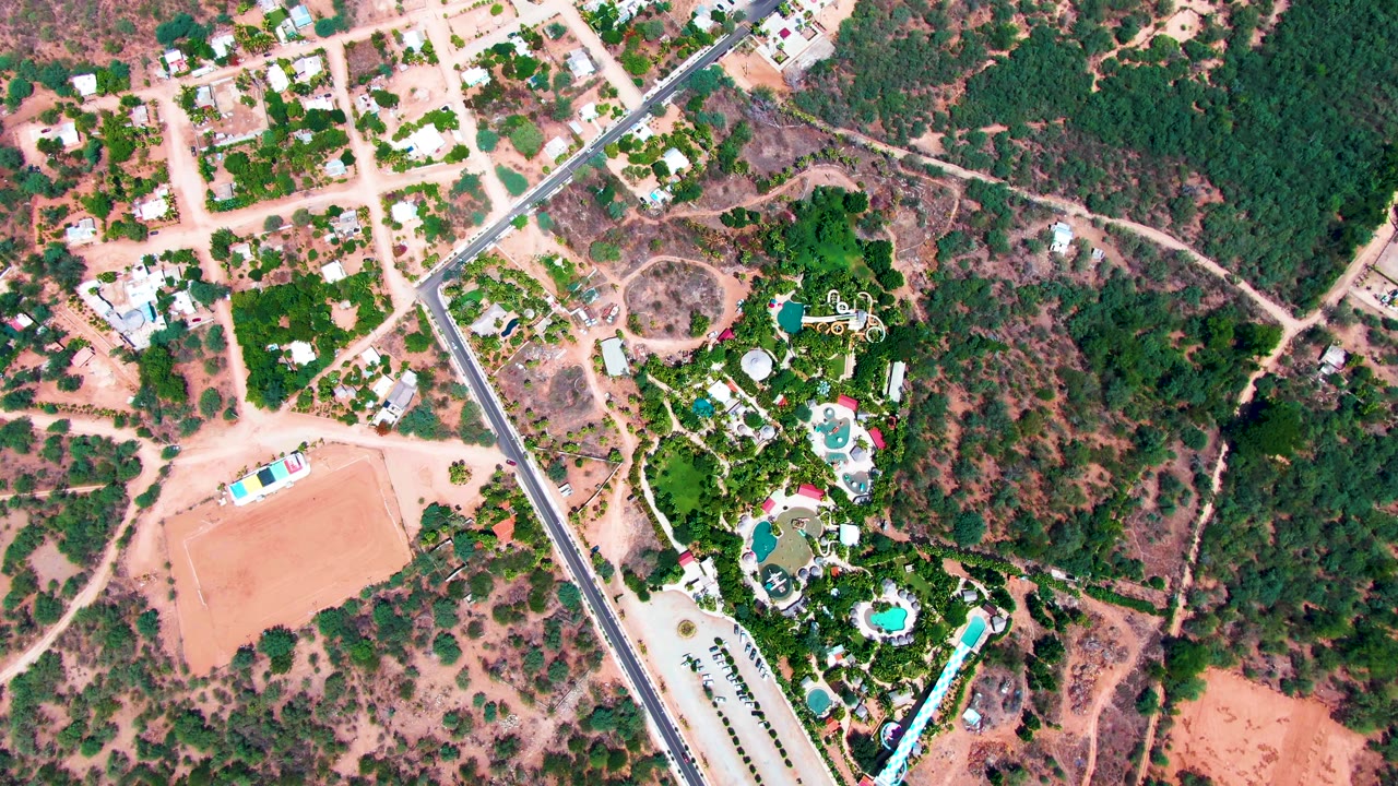 High overhead view in Caduano, Baja California Sur, showing a diagonal road beside homes, blue pools, green trees, and dry reddish terrain with no visible people.