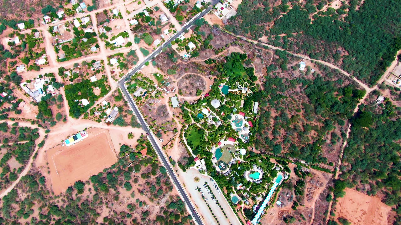 Drone-like overhead frame in Caduano with the road cutting past a green property, several blue pools, pale dirt tracks, and dry open ground with no visible people.