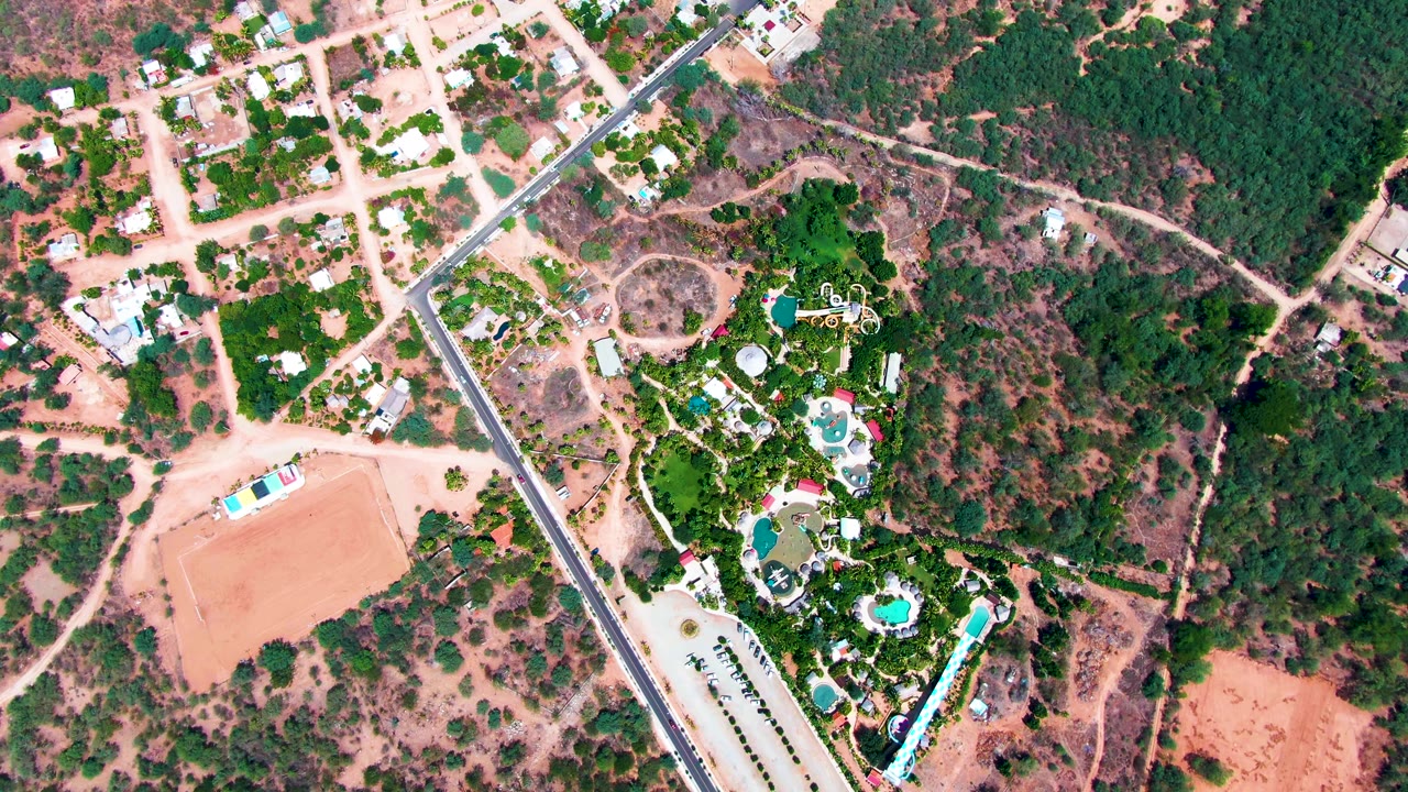 Overhead Caduano frame showing the road along the left side of a green pool property, with dry lots, houses, dirt paths, and no visible people.