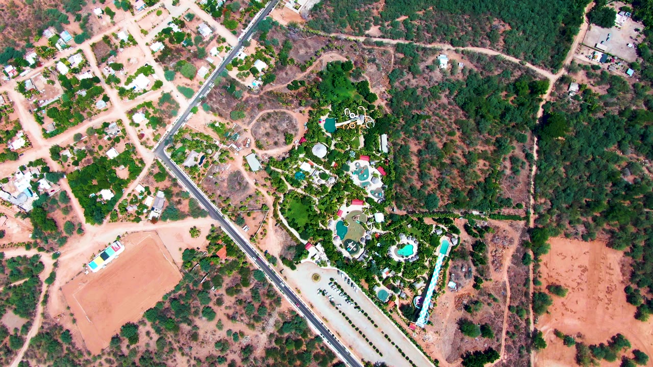 Overhead drone frame in Caduano, Baja California Sur showing a diagonal paved road, pale dirt blocks, scattered homes, dense trees, and blue pools, with no visible people.
