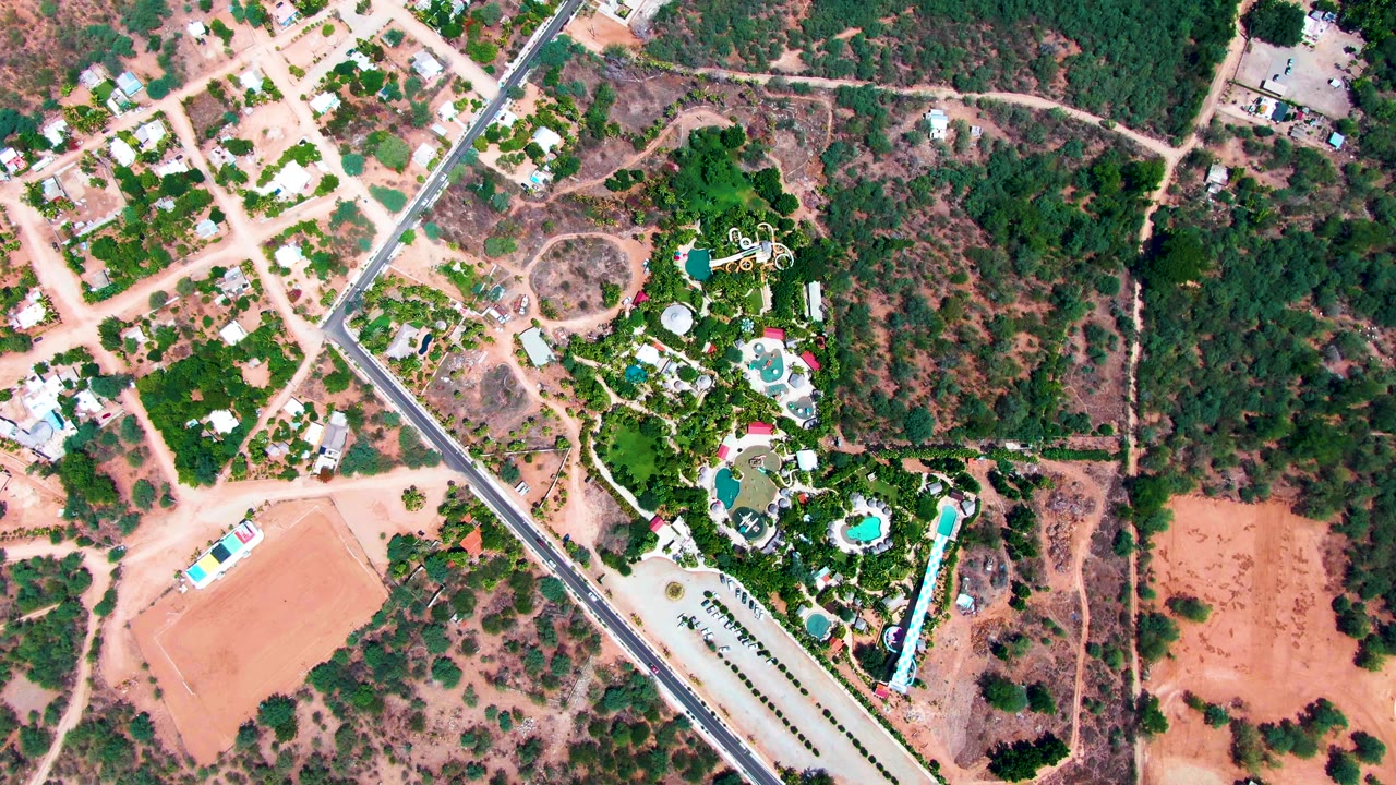 Aerial frame over Caduano with the paved road running diagonally past dry residential blocks and a greener pool area, with no visible people.