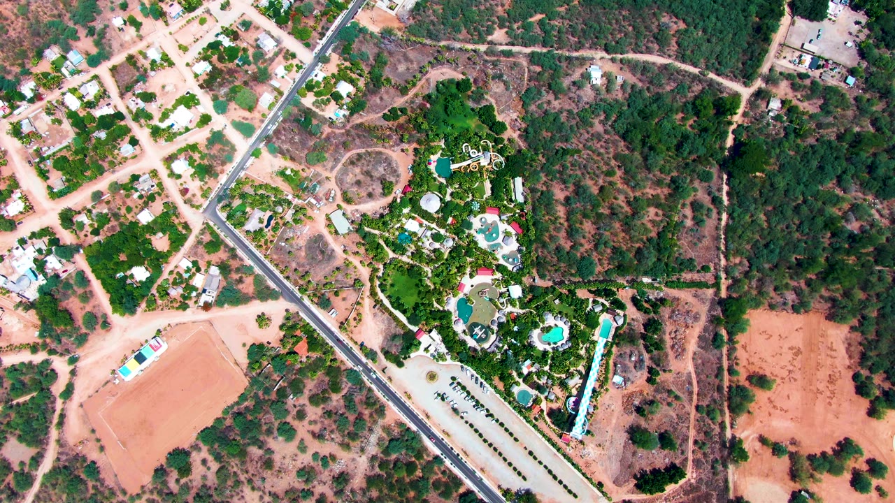 Overhead view in Caduano showing houses clustered near pale dirt roads, a diagonal paved road, scrubby open ground, tree cover, and bright pool water, with no visible people.