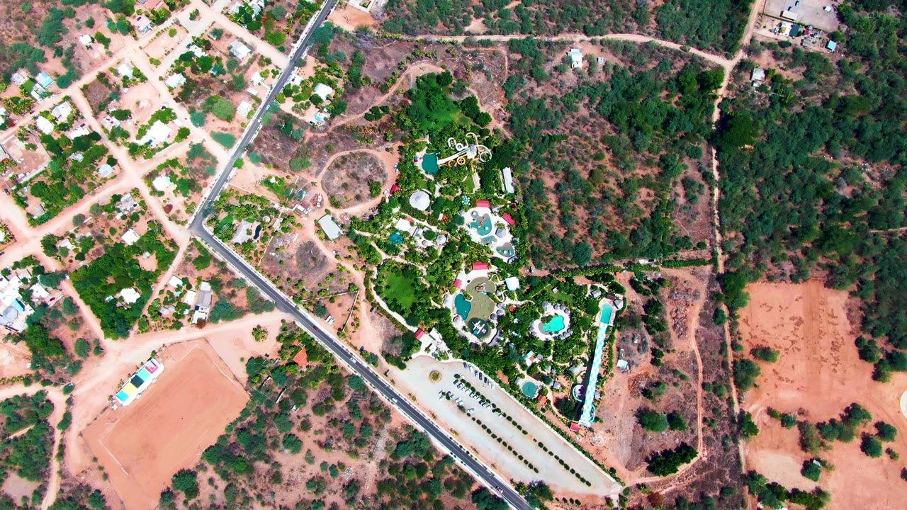 Overhead frame in Caduano, Baja California Sur, showing a diagonal paved road beside dry reddish lots, scattered homes, green resort landscaping, turquoise pools, parking, and no visible people.