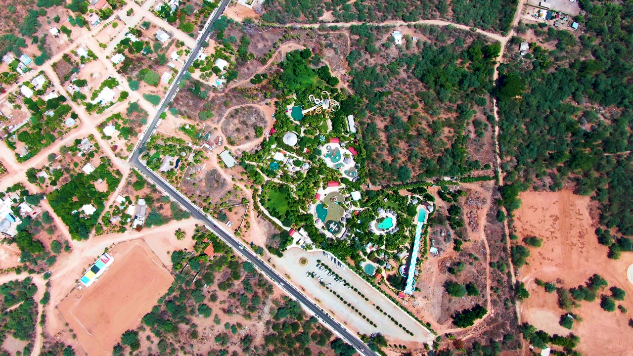 Aerial frame in Caduano, Baja California Sur, with the paved road still crossing diagonally past resort pools, green trees, pale dirt paths, open scrubland, and no visible people.